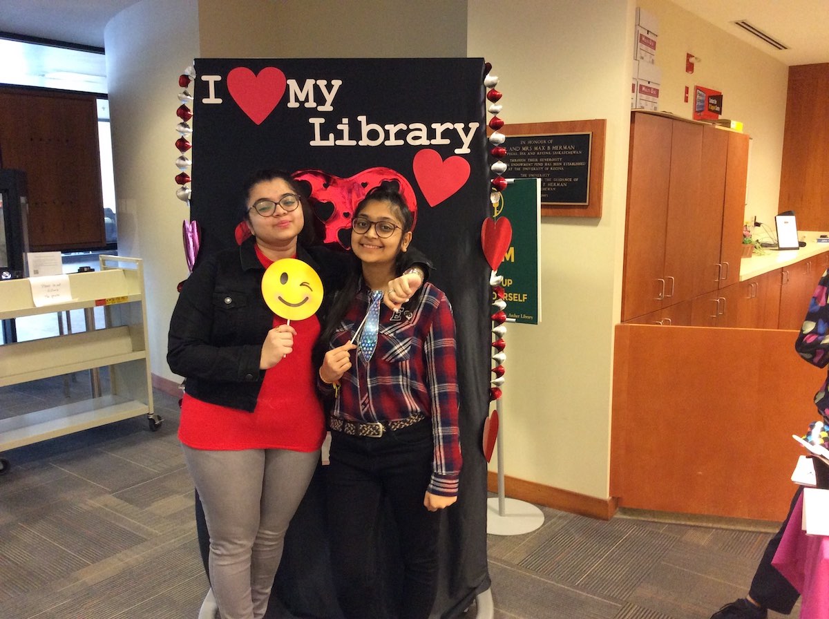 Two smiling students pose in front of the I love my Library backdrop. The one on the left is holding a winking smiley face prop on a stick and has her arm around the other who is holding up a prop necktie on a stick.
